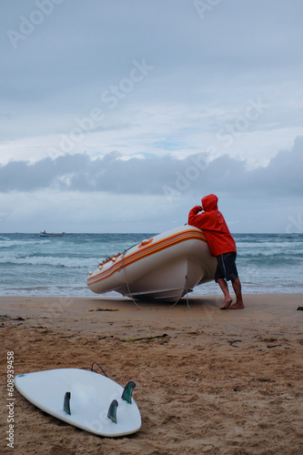 Surfboard on a beach with a man leaning on an inflatable raft and a stormy sky.