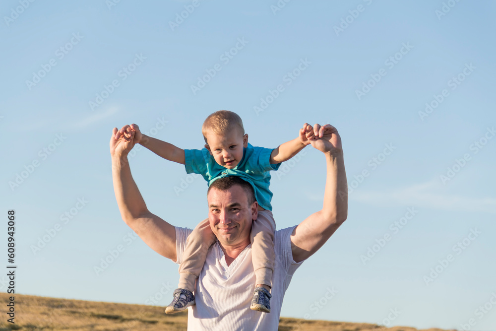 Dad and his little son are having fun walking in a field with ripe wheat. The child is sitting on the shoulders of the father. Dad and his little son are having fun walking in a field with ripe wheat. The child is sitting on the shoulders of the father.