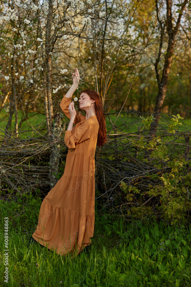 an elegant, sophisticated woman poses standing near a wicker fence in a dacha in a long orange dress, raising her hands up