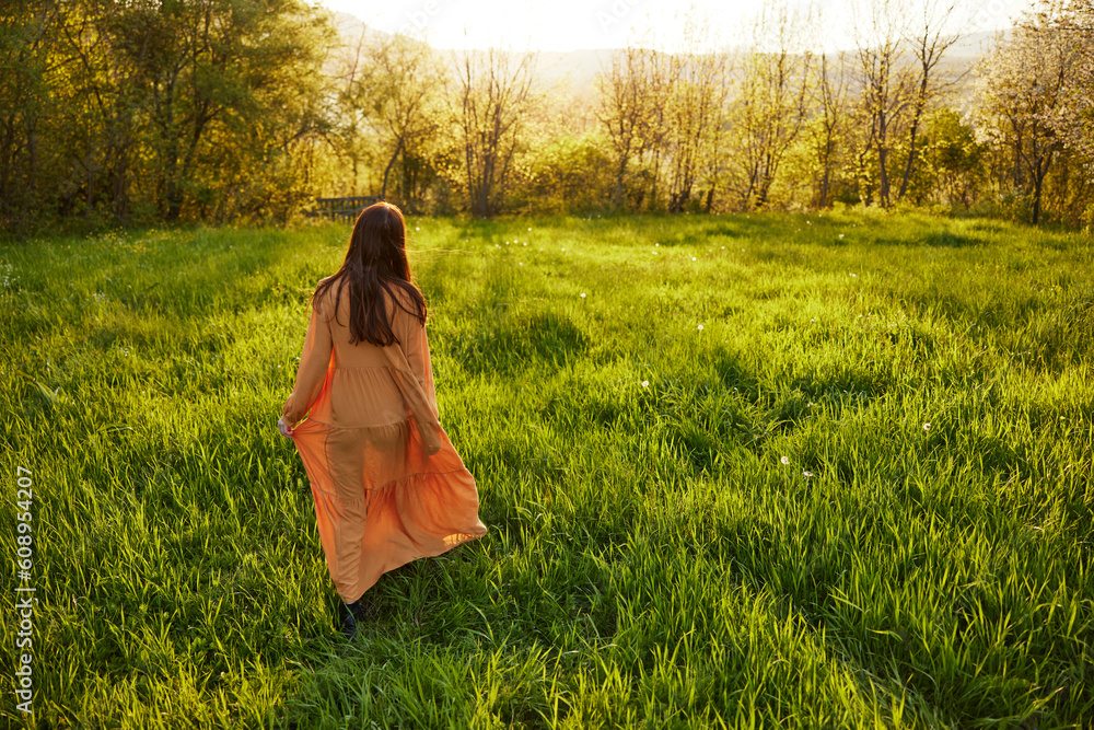 an attractive, slender, red-haired woman walks through a field during sunset, in a long orange dress enjoying unity with nature and relaxation holding the edge of her clothes an attractive, slender, red-haired woman walks through a field during sunset, in a long orange dress enjoying unity with nature and relaxation holding the edge of her clothes