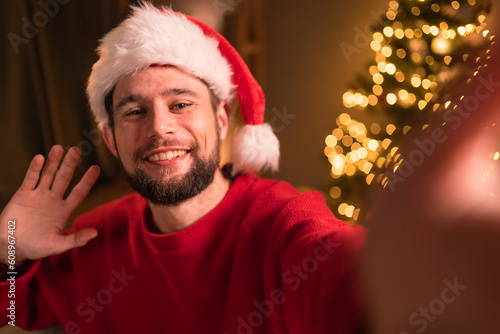 Happy bearded man in Santa Claus hat holding phone waving hand taking selfie, video calling, recording video Merry Christmas greeting or shooting vlog photo