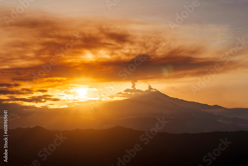 mysterious landscape of great erupting volcano with smoke from craters and snow on slopes in orange light of sunset. eruption of vulcan © Yaroslav