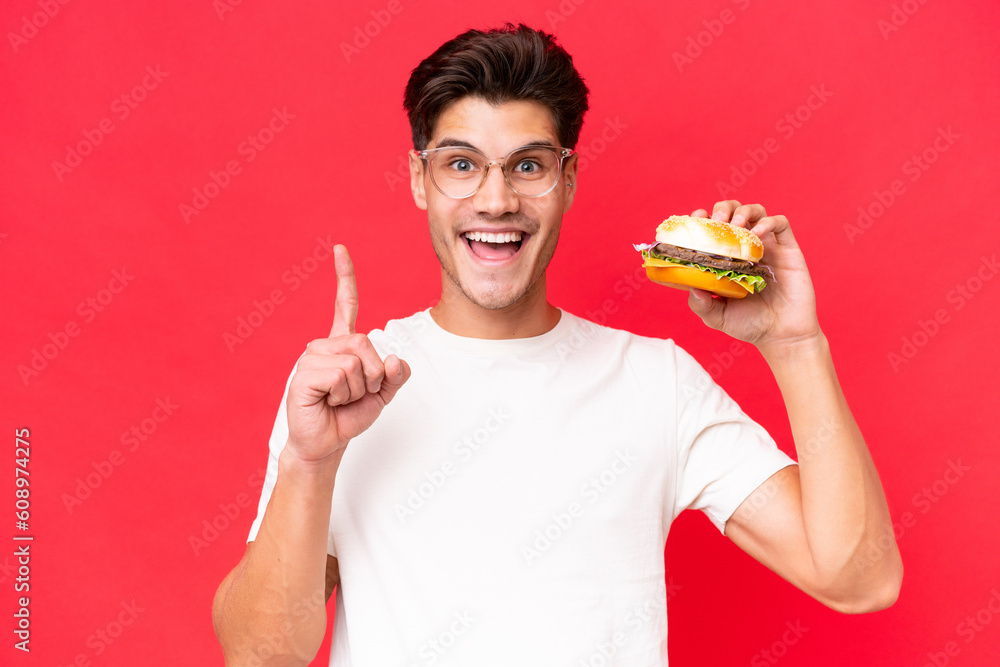 Young Caucasian man holding a burger isolated on red background pointing up a great idea