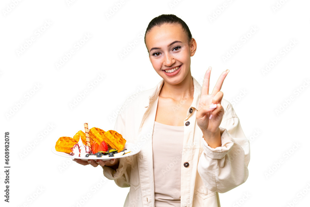Young Arab woman holding waffles over isolated background smiling and showing victory sign Young Arab woman holding waffles over isolated background smiling and showing victory sign