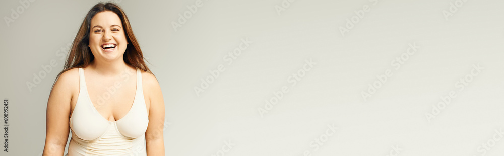 Obraz premium portrait of happy and curvy woman with plus size body posing in beige bodysuit while laughing on grey background, body positive, figure type, looking at camera while standing in studio, banner