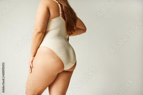 cropped view of curvy woman wearing beige bodysuit and standing with hand on hip isolated on grey background, self-confidence, figure type, body positivity movement