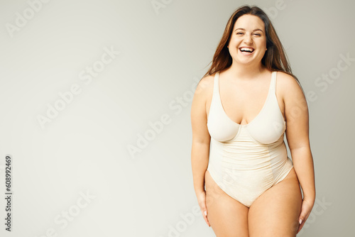 portrait of happy and curvy woman with plus size body posing in beige bodysuit while laughing on grey background, body positive, figure type, looking at camera while standing in studio