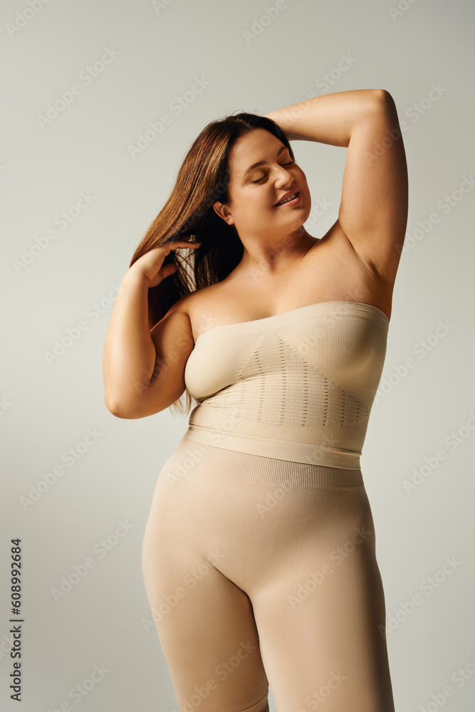 Fototapeta premium radiant woman with closed eyes touching hair and posing with bare shoulders in underwear isolated on grey background in studio, body positive, self-love, happy plus size