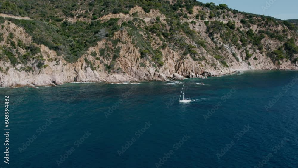 Side shot of idyllic white sail boat drift on motor power next to shore towards private cove or port. Summer spanish vacation in Costa Brava or Mallorca Side shot of idyllic white sail boat drift on motor power next to shore towards private cove or port. Summer spanish vacation in Costa Brava or Mallorca