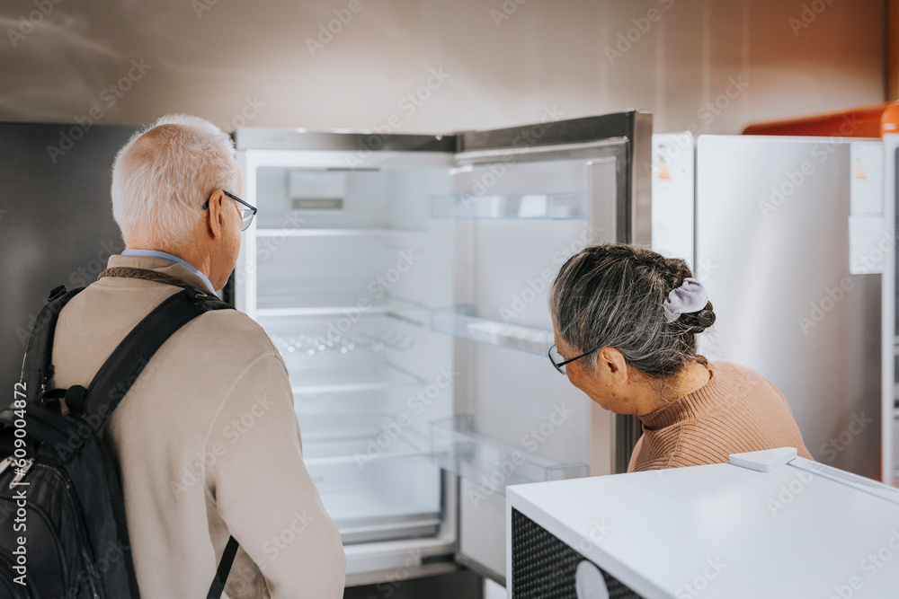 Senior couple opening and examining refrigerator while shopping in appliances store Senior couple opening and examining refrigerator while shopping in appliances store