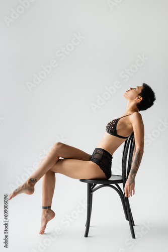 side view of graceful woman with short brunette hair and sexy tattooed body sitting on chair in black bra with pearl beads and lace panties on grey background, erotic photography