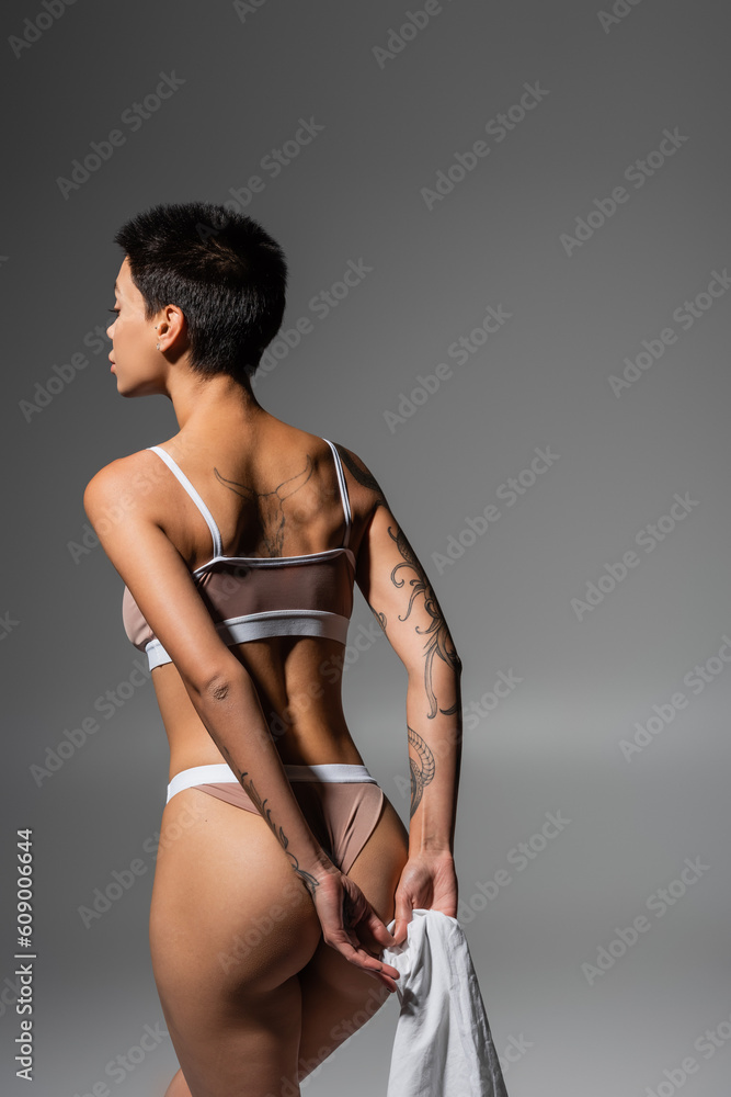 young and desirable woman with short brunette hair and sexy tattooed body  holding white bed sheet while posing in beige lingerie in studio on grey  background, erotic photography Stock Photo