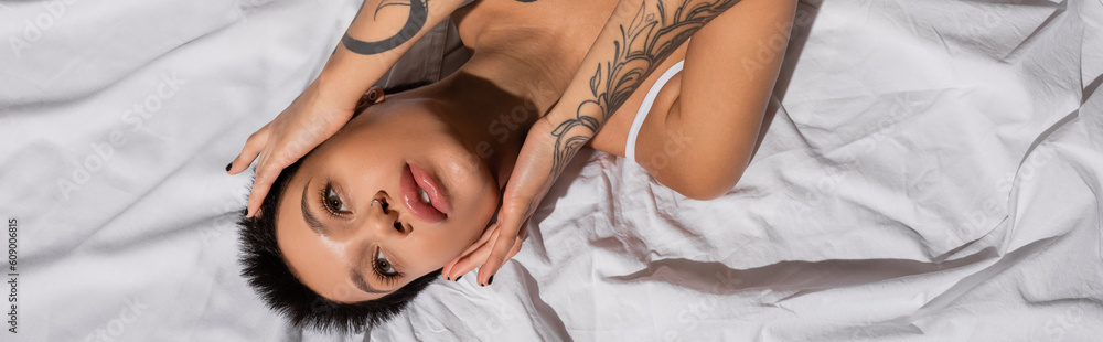 top view of young, tattooed and charming woman with short brunette hair touching face and looking away while laying on white bedding in studio, erotic photography, banner