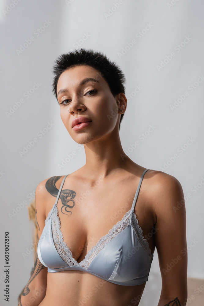 portrait of young ad sexy woman in silk bra, with tattooed body, short  brunette hair and natural makeup looking at camera while posing in bedroom  at home on blurred background Photos