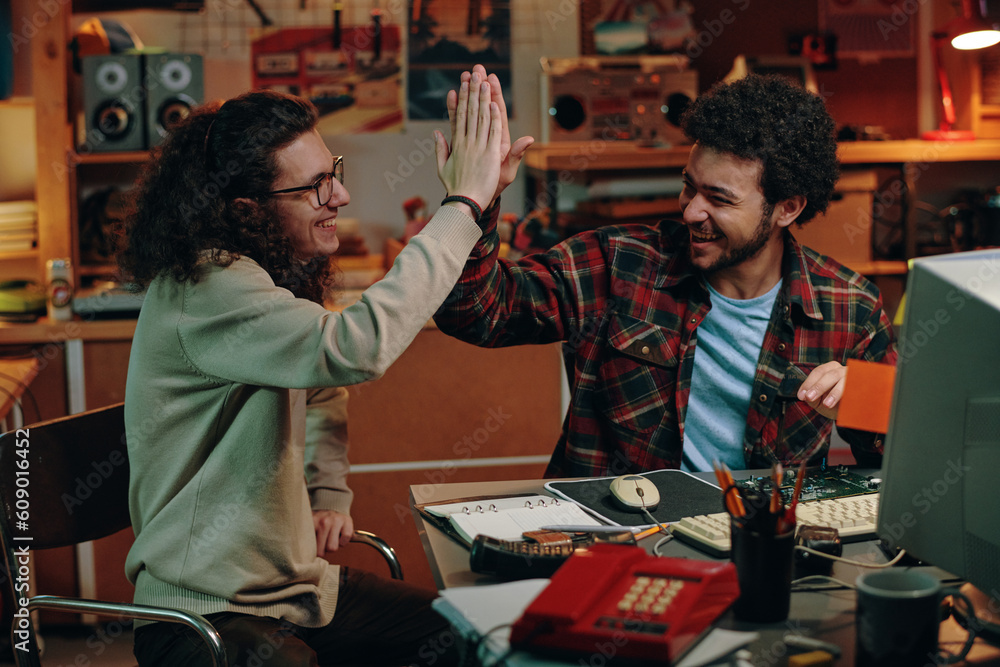 Obraz premium Young people giving high-five to each other while sitting at table in workshop, they happy their successful project
