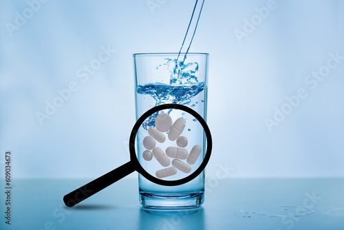 A magnifying glass with large white tablets in front of a glass of water. Concept: antibiotics in drinking water