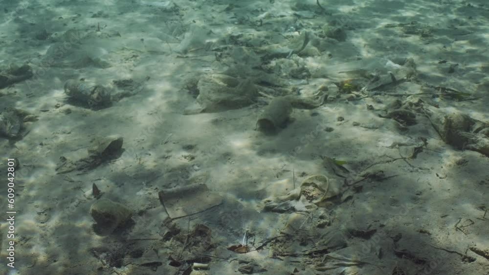 Plastic garbage lies on sandy bottom of sea in shallow water on bright sunny day in sun glare. Camera moving forwards above seabed polluted with plastic and other debris, Slow motion Plastic garbage lies on sandy bottom of sea in shallow water on bright sunny day in sun glare. Camera moving forwards above seabed polluted with plastic and other debris, Slow motion