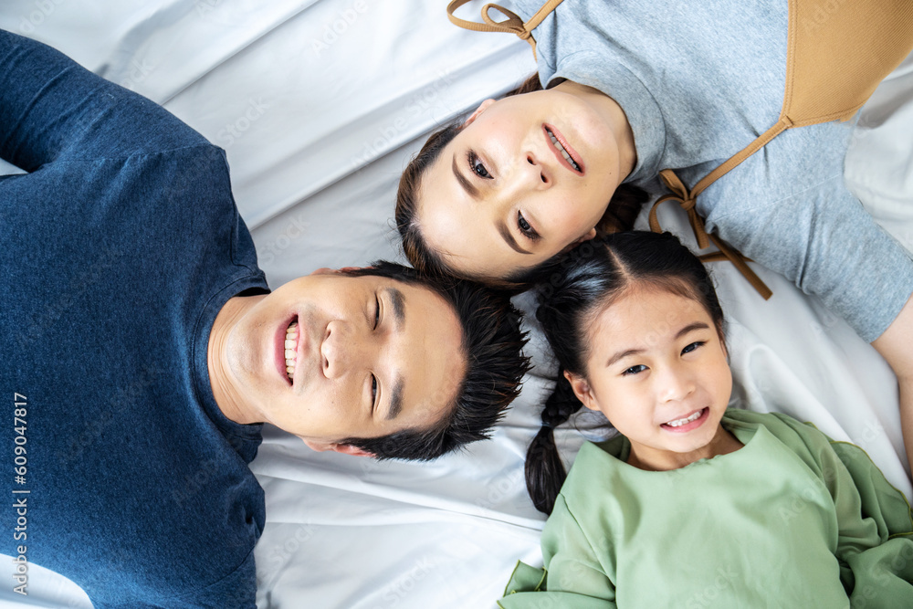 Top view of Happy Asian beautiful young mother, father and their daughter looking at camera and smiling while lying on bed head to head in bedroom