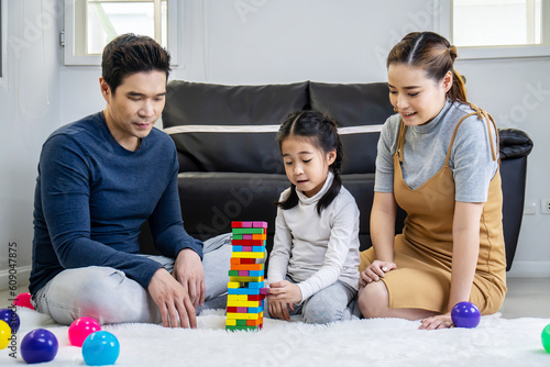 Happy asian family father and mother with little asian girl smiling playing with building tower from wooden blocks, taking tiles in turn from underneath until it falls, in moments good time at home