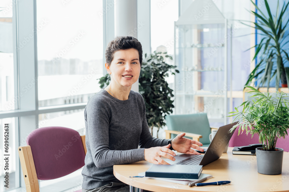 Neutral gender middle aged woman with no make-up in casual clothing using laptop while working indoors on workstation in an open space office, coworking, library. Remote workplace, selective focus. Neutral gender middle aged woman with no make-up in casual clothing using laptop while working indoors on workstation in an open space office, coworking, library. Remote workplace, selective focus.