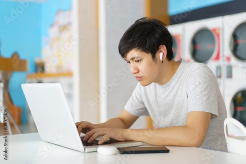 Asian man using laptop or notebook computer for online video conference and entertainment while waiting for his clothes wash in the self-service automatic washing or laundry machine. photo