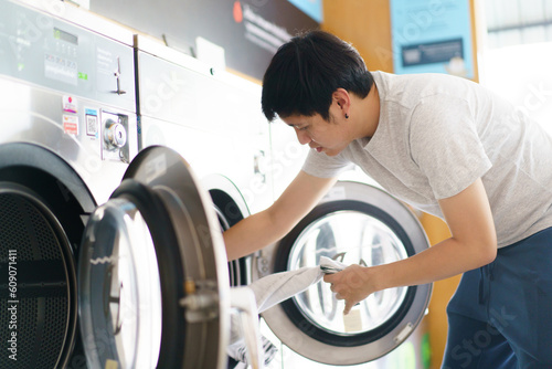 Canvas Print Asian man putting an used or dirty clothes in the self-service automatic laundry washing machine, Asian man using smartphone to access self service KIOS laundry machine