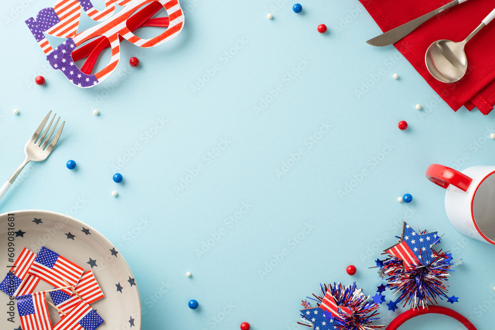 Fototapeta premium Show your American spirit with this table arrangement. Top view, including plate, cup, cutlery, napkin, sprinkles, headband, glasses, national flag on backdrop of pastel blue. Frame for text or promo