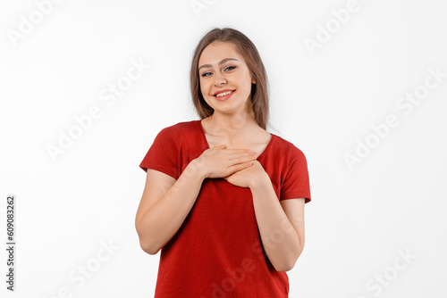 Happy thankful young woman holding hands on chest, feeling gratitude, mental health balance, no stress, peace of mind concept. Winsome female model in red t shirt posing over white studio background