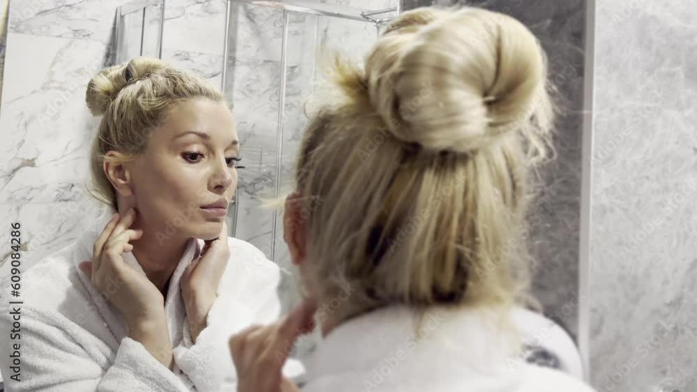 mid age adult blonde woman wears bathrobe and towel in bathroom applying rejuvenating tightening antiage face skin care cream treatment. Daily beauty routine. mid age adult blonde woman wears bathrobe and towel in bathroom applying rejuvenating tightening antiage face skin care cream treatment. Daily beauty routine.