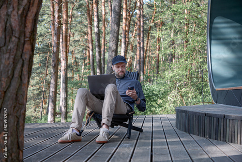 a young man with a beard and in a cap sits in nature against the backdrop of a pine forest and works in a laptop with a phone in his hands