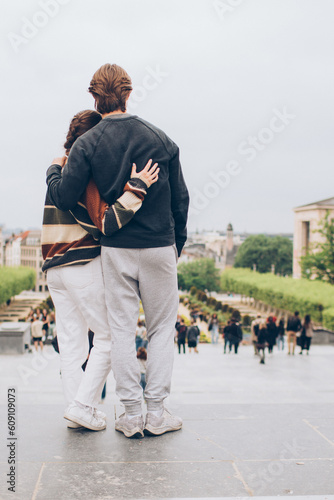 couple standing on the top of a staircase looking down at a crowd of people in a city in europe embracing with a camera view of the backs wearing sweatshirts