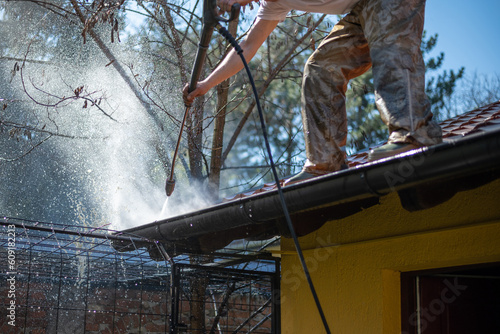 Worker standing on the roof and cleaning rain gutter with high pressure water jet. Professional using equipment for roof gutter cleaning. Maintenance and housekeeping concept of drainage channel. 