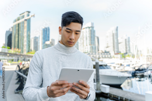 Portrait of handsome asian businessman  holding digital tablet, watching video, check email, working online standing on street. Serious modern Chinese man reading e book outdoors. Technology concept © Maria Vitkovska