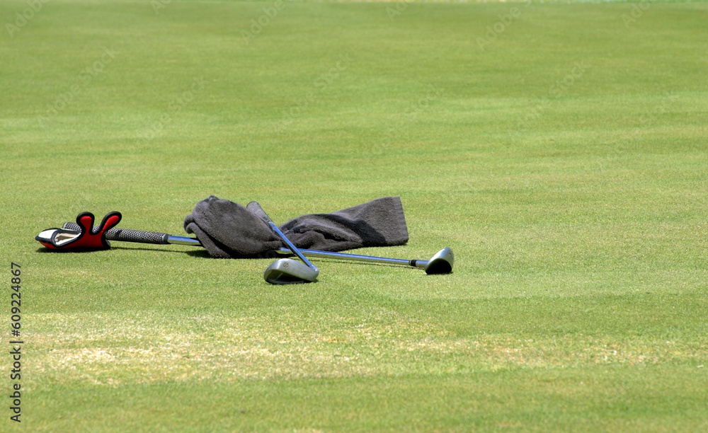 Obraz premium Wet towel and two golf clubs lying on the green grass at the golf course. Sunny day.