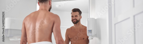 Happy bearded man with bare chest wrapped with towel looks in mirror in modern barthroom photo