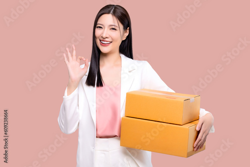 Portrait of young Asian business woman with boxes showing OK sign, carry delivery goods, standing isolated over pink background.