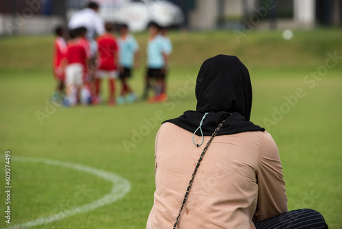 Moms sitting and watching their sons playing football in a school tournament on a sideline with a sunny day. Sport, outdoor active, lifestyle, happy family and soccer mom and soccer dad concepts.