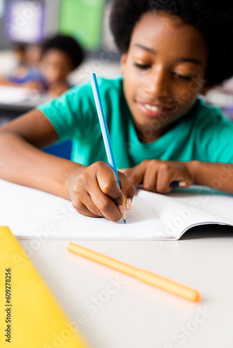 Portrait of happy african american elementary schoolboy writing in notebook at desk in class