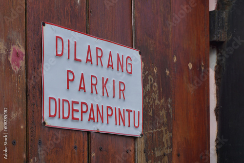 Sign "No parking in front of the door" in Bahasa. Mounted in front of a brown wooden door.