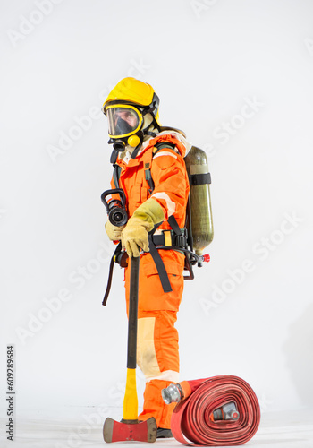 Side view of firefighter stands on a white background using an iron axe on the ground while holding a fire hose in one hand and an oxygen tank behind his back with the other and looking to the camera.