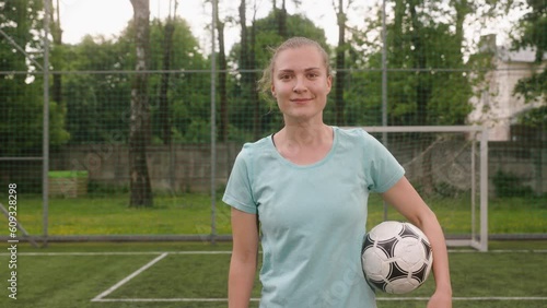 A portrait of a female football player training on a mini-football stadium. The girl holds a ball in her hand, looks into the camera, and smiles.
