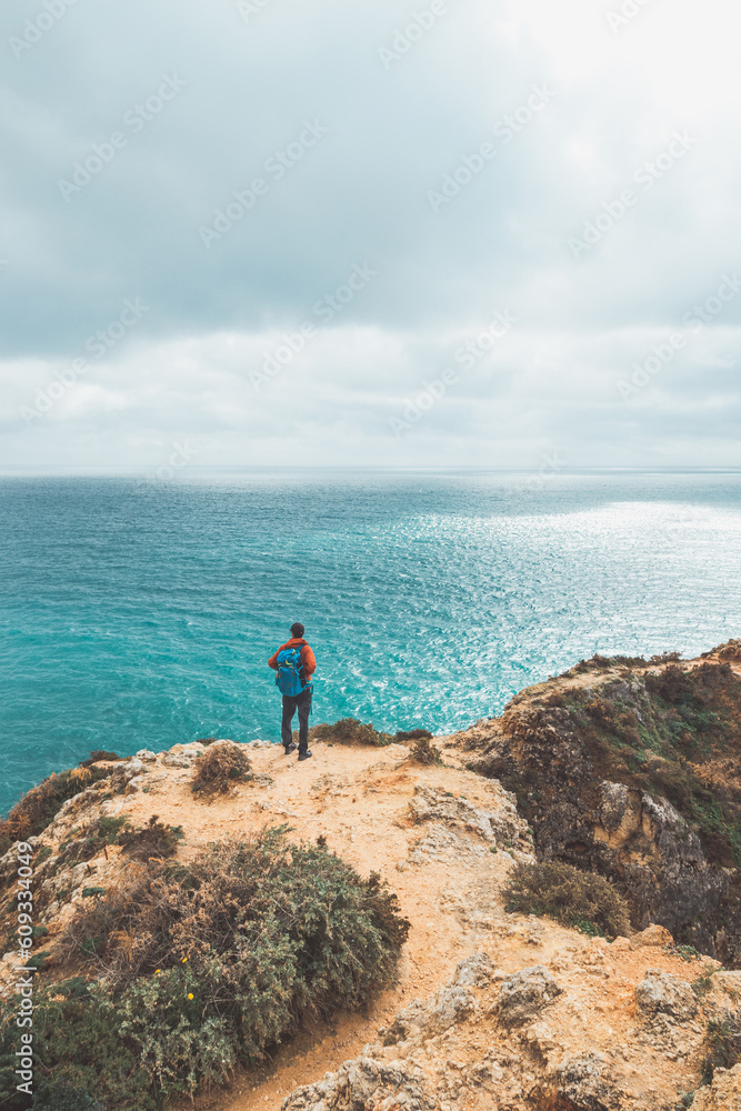Adventurer with a backpack stands on the edge of a cliff and observes the beautiful group of yellow-gold rocks of Punta de la Piedad in Lagos, in the Algarve region of southern Portugal