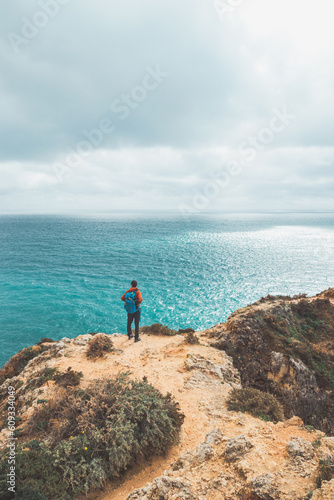 Adventurer with a backpack stands on the edge of a cliff and observes the beautiful group of yellow-gold rocks of Punta de la Piedad in Lagos  in the Algarve region of southern Portugal