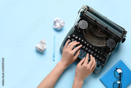Fototapeta Naklejka Na Ścianę i Meble -  Woman typing on typewriter with crumpled paper, notebook and eyeglasses on light blue background