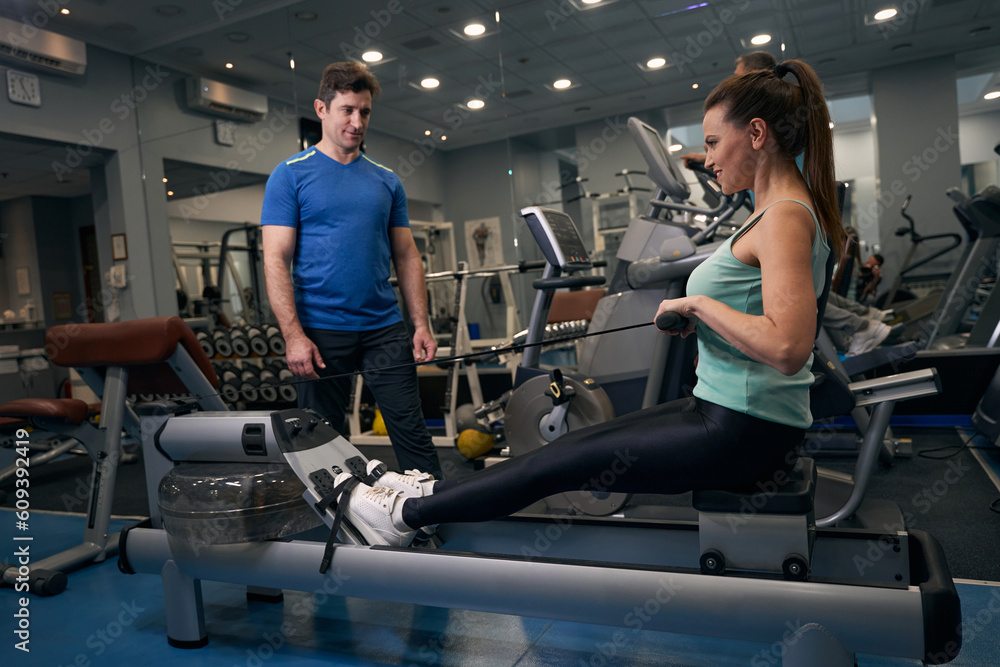 Female performing upper back exercise supervised by personal trainer Female performing upper back exercise supervised by personal trainer