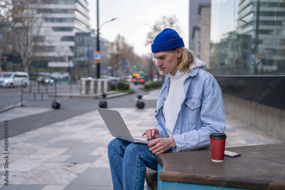 Focused young man 20s wearing denim clothes and beanie hat sit on bench with coffee cup working online on laptop. Guy student sits on city street studying online. Urban lifestyle and freelancing Focused young man 20s wearing denim clothes and beanie hat sit on bench with coffee cup working online on laptop. Guy student sits on city street studying online. Urban lifestyle and freelancing