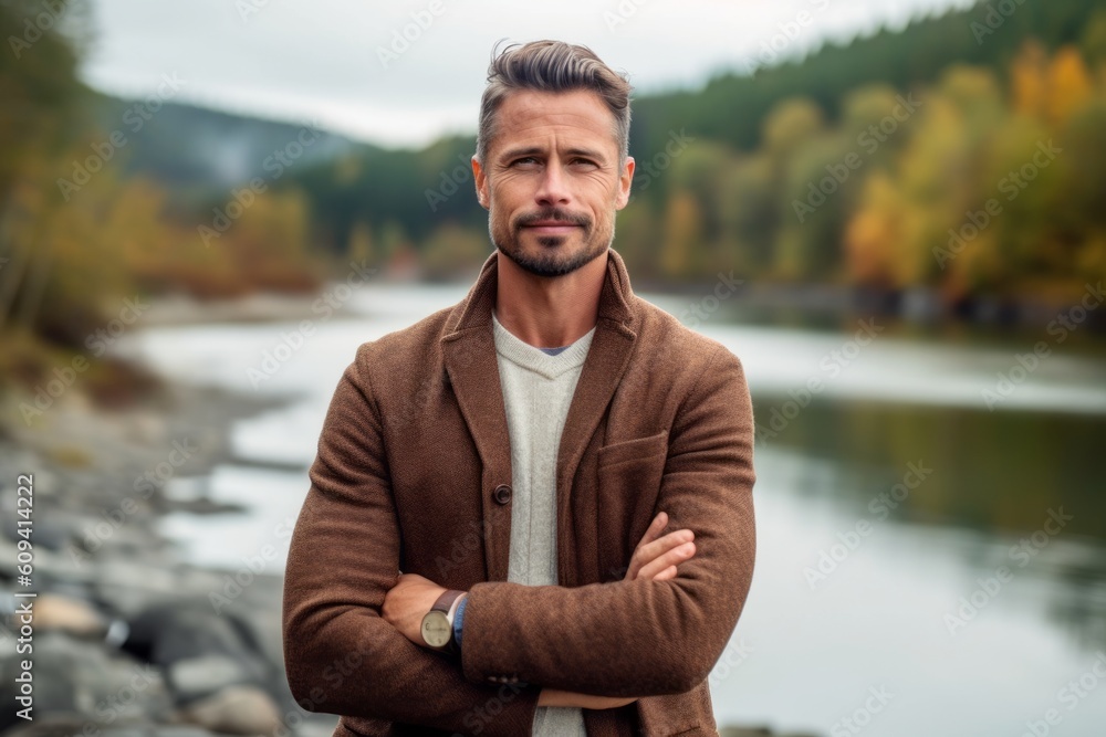 Fototapeta premium Handsome man in a brown coat with crossed arms on the background of a mountain river