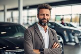 Handsome man in suit is standing with crossed arms in car dealership.