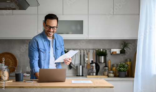 Smiling male financial advisor holding income tax documents and using laptop on counter in kitchen. Happy young freelancer analyzing investment report over wireless computer in home office