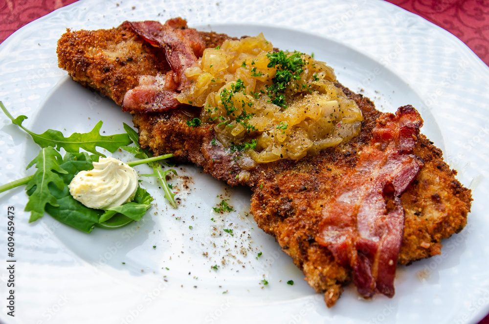 White plate with a large schnitzel with onions, bacon, rucola, cream and parsley in a restaurant terrace in Germany on a sunny day